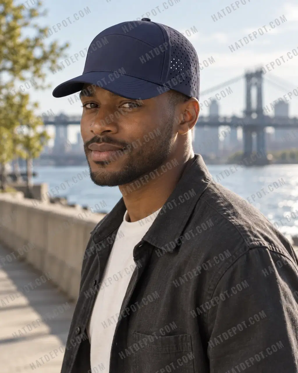 Model beside a sunlit riverside walkway in the navy Six-Panel Perforated Snapback, showing its seamless front, curved brim and vented sides.